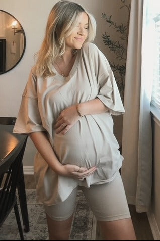 woman wearing a beige maternity top and shorts, holding her baby bump while standing by a window