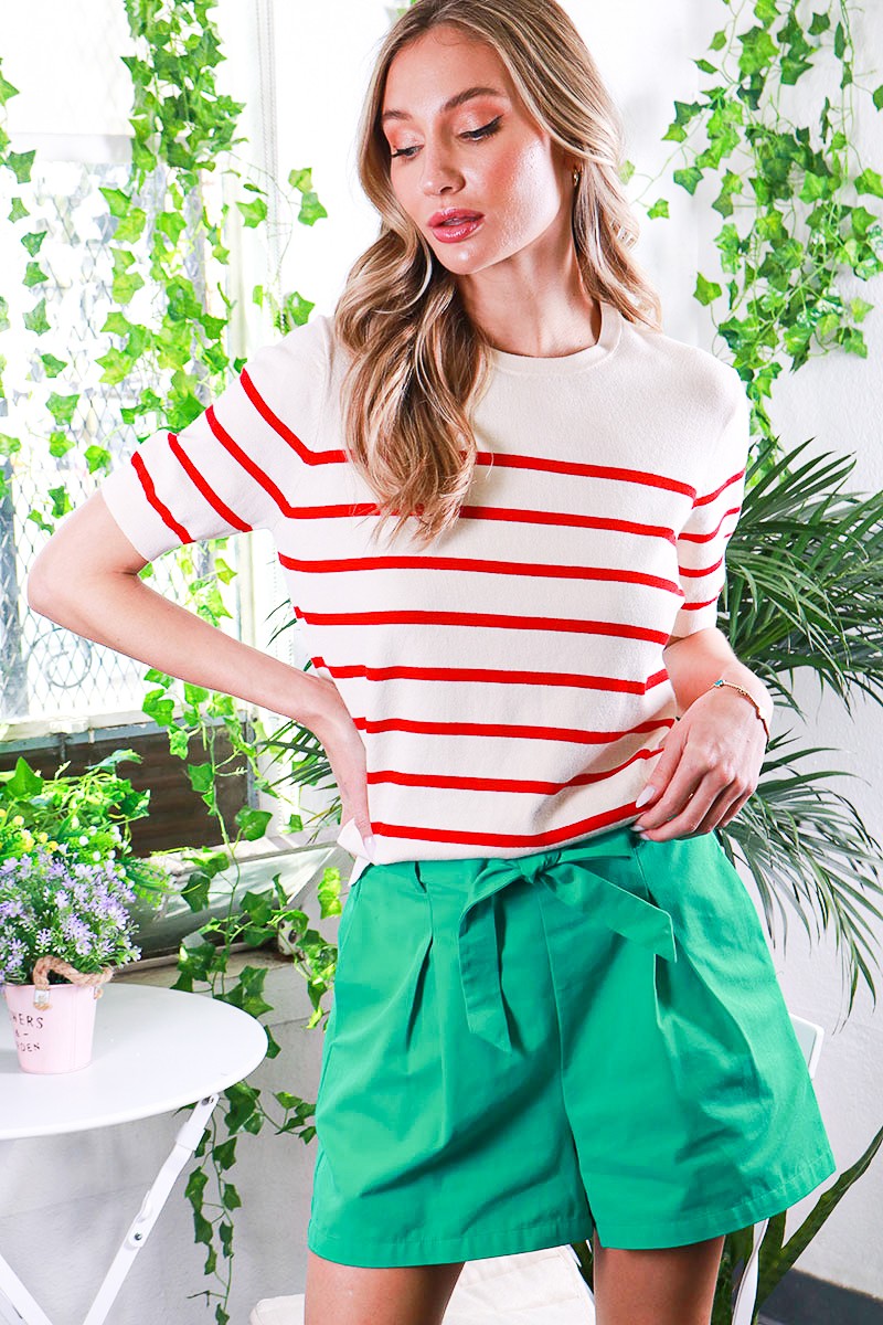 woman wearing a cream striped sweater with red stripes and green high-waisted shorts, posing in a vibrant indoor setting