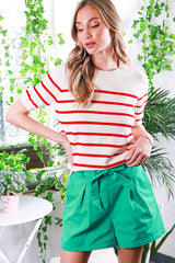 woman wearing a cream striped sweater with red stripes and green high-waisted shorts, posing in a vibrant indoor setting
