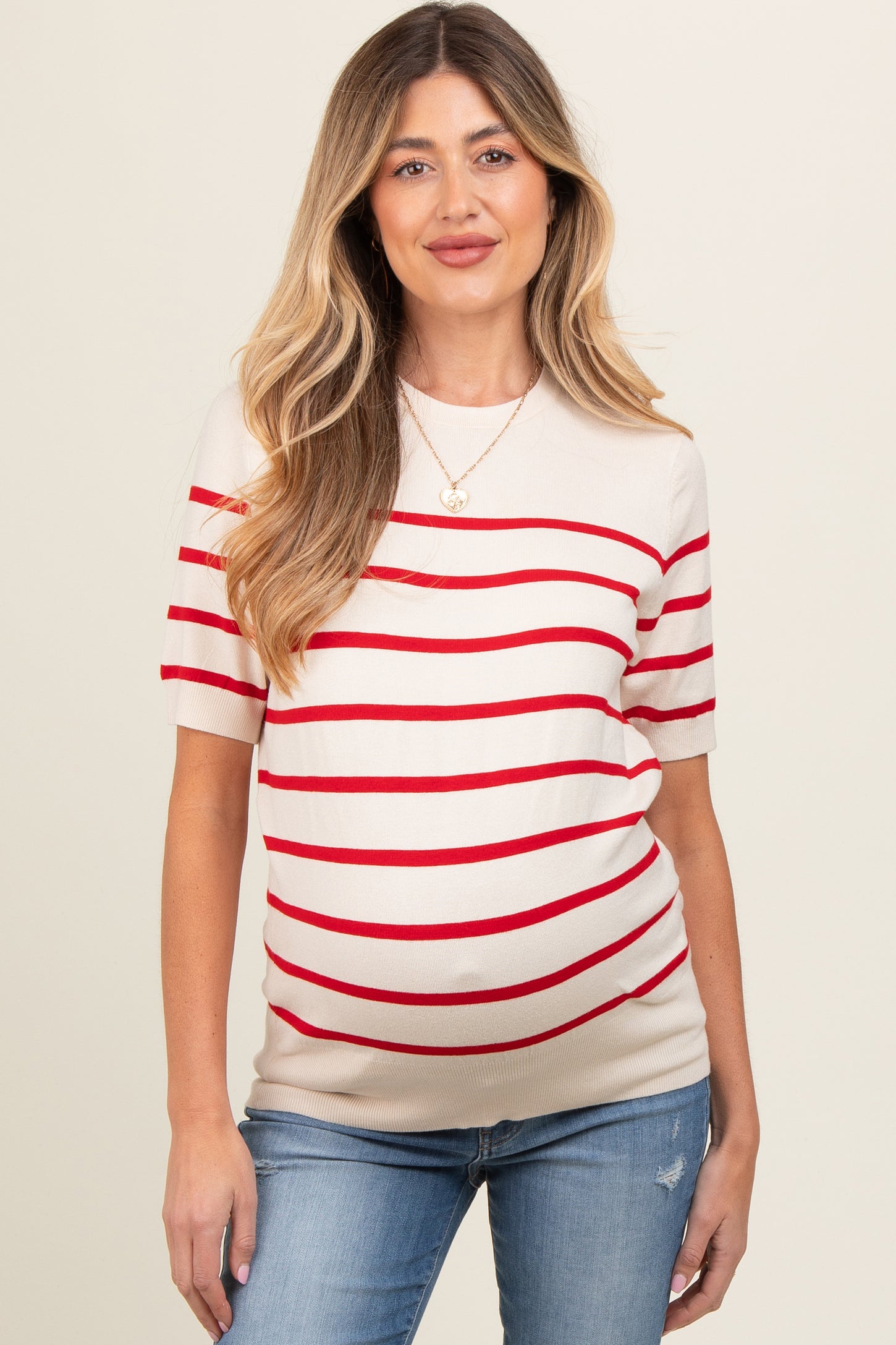 woman wearing a beige maternity top with red stripes and a visible baby bump, paired with blue jeans