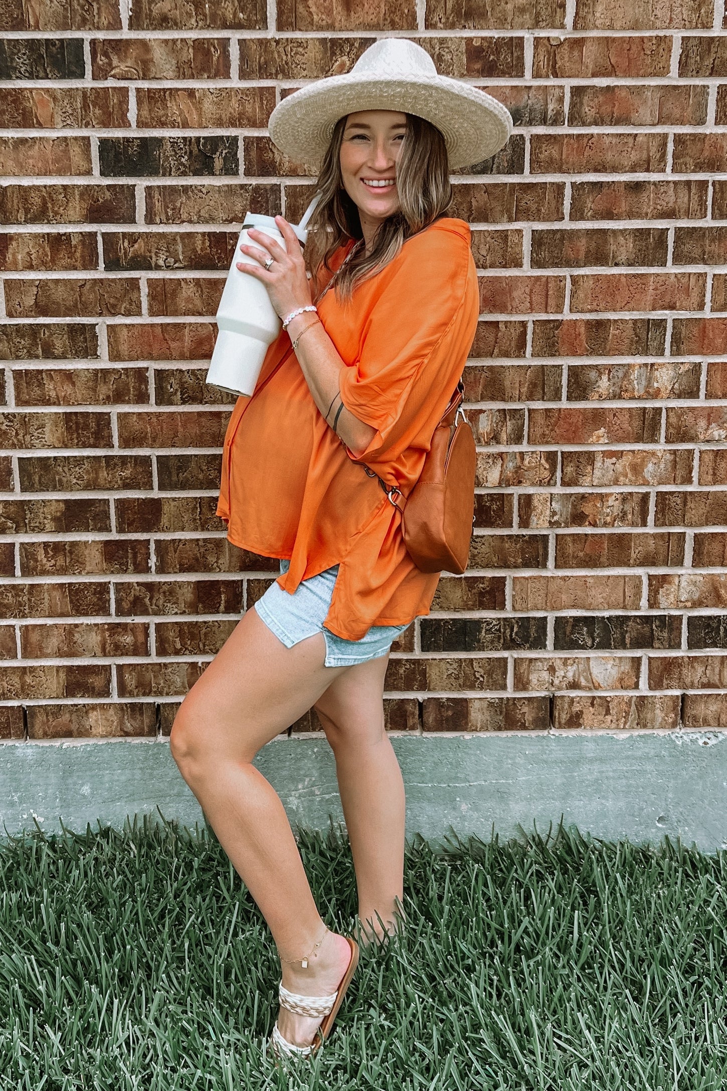 woman wearing an orange maternity top and light blue shorts, holding a drink and posing with a straw hat and handbag