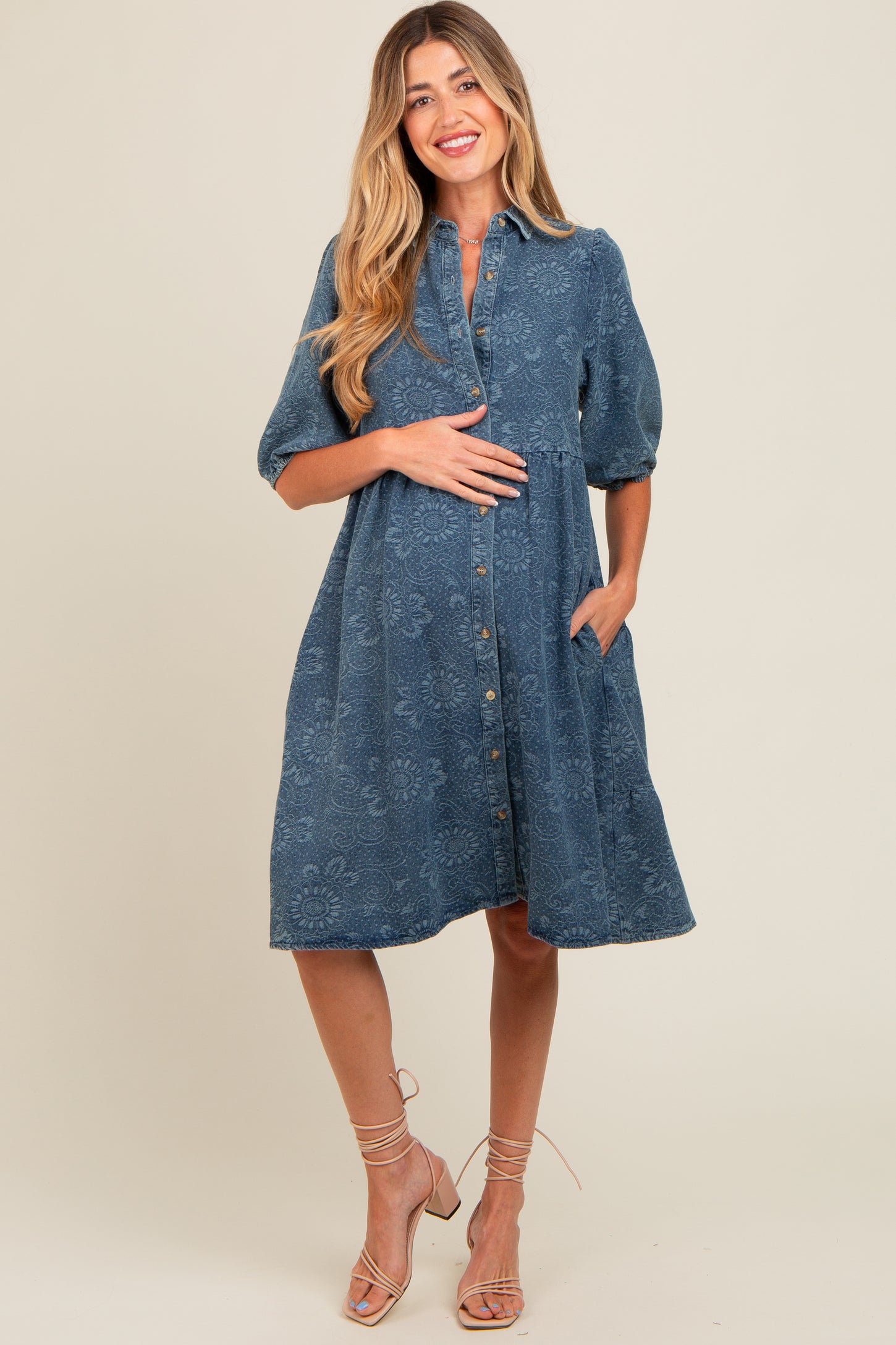 woman wearing a denim blue maternity dress with short puff sleeves, button front, and side pockets, showcasing her baby bump