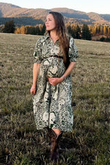 woman wearing a green and white patterned maternity dress and holding her baby bump in a field