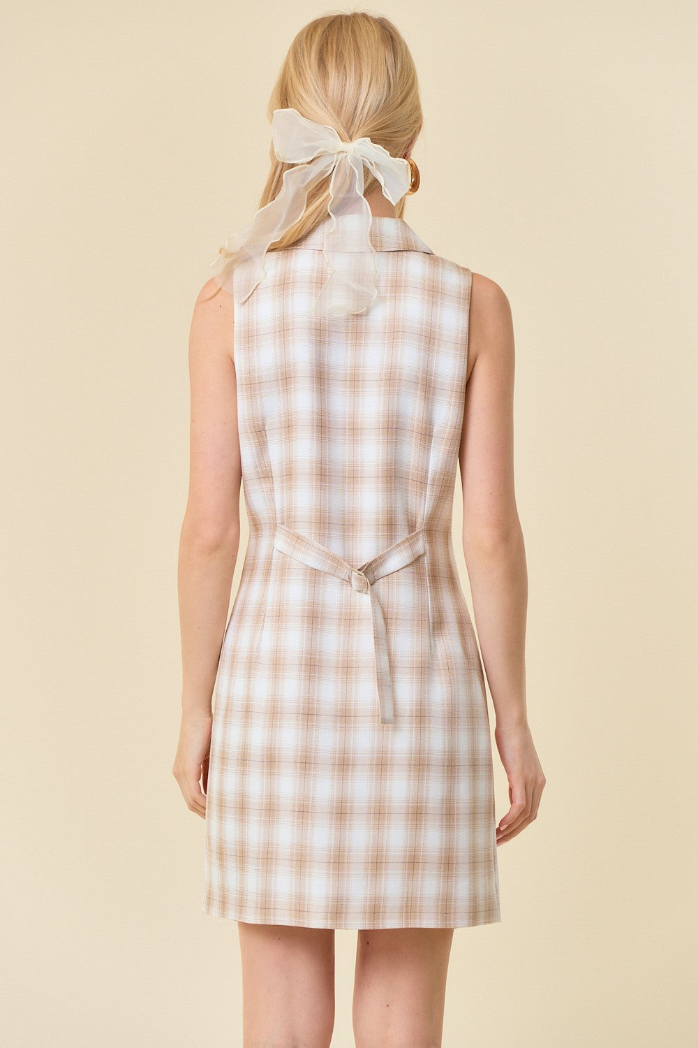 woman wearing a beige and cream plaid sleeveless dress with a tie waist and a sheer hair ribbon