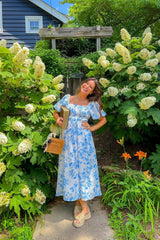 woman wearing a white and blue floral maxi dress with puff sleeves, standing in front of lush greenery and flowers