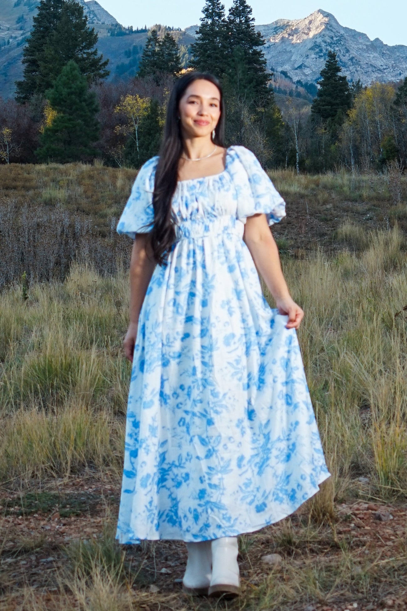 woman wearing a long white dress with blue floral patterns and puff sleeves, standing in a grassy field with mountains in the background