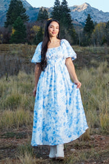 woman wearing a long white dress with blue floral patterns and puff sleeves, standing in a grassy field with mountains in the background