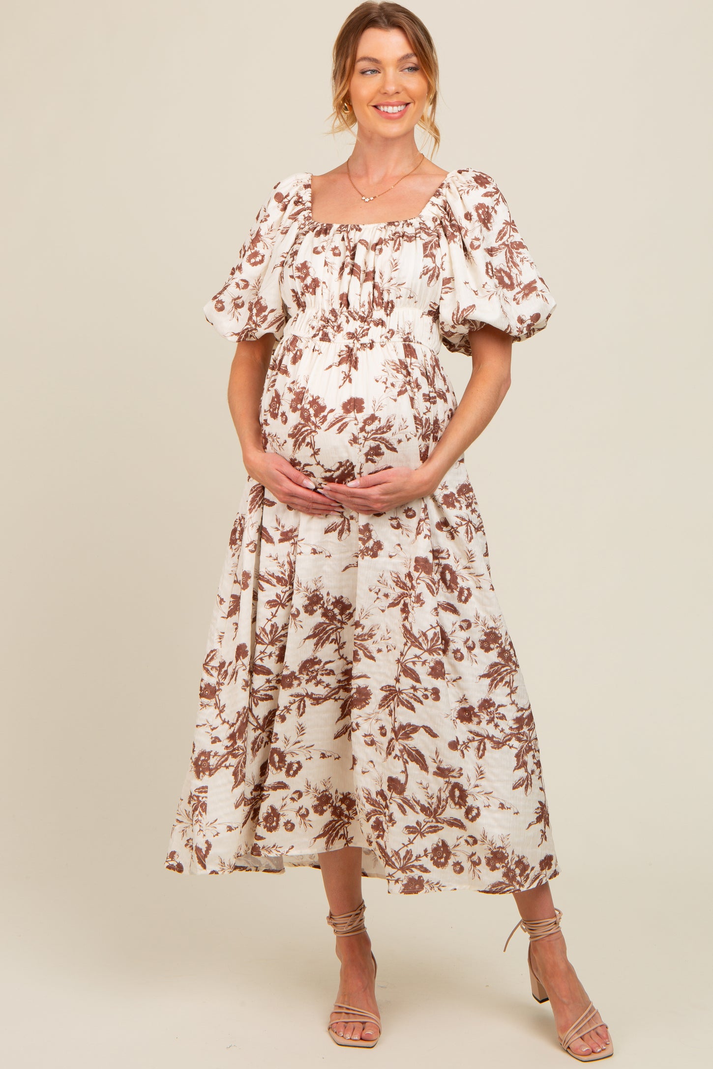 woman wearing a cream floral print maternity dress with puff sleeves and a gathered bust, holding her baby bump