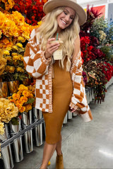 woman wearing a brown and white checkered cardigan over a mustard ribbed midi dress, holding a drink and smiling in a floral setting