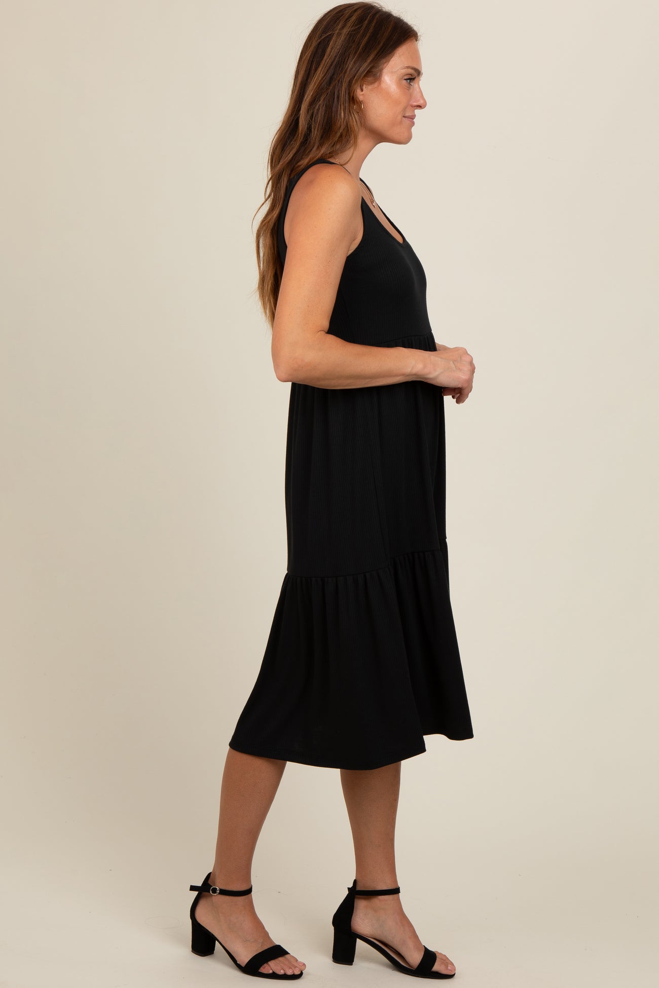 woman wearing a black sleeveless midi dress with a tiered skirt and round neckline, standing sideways