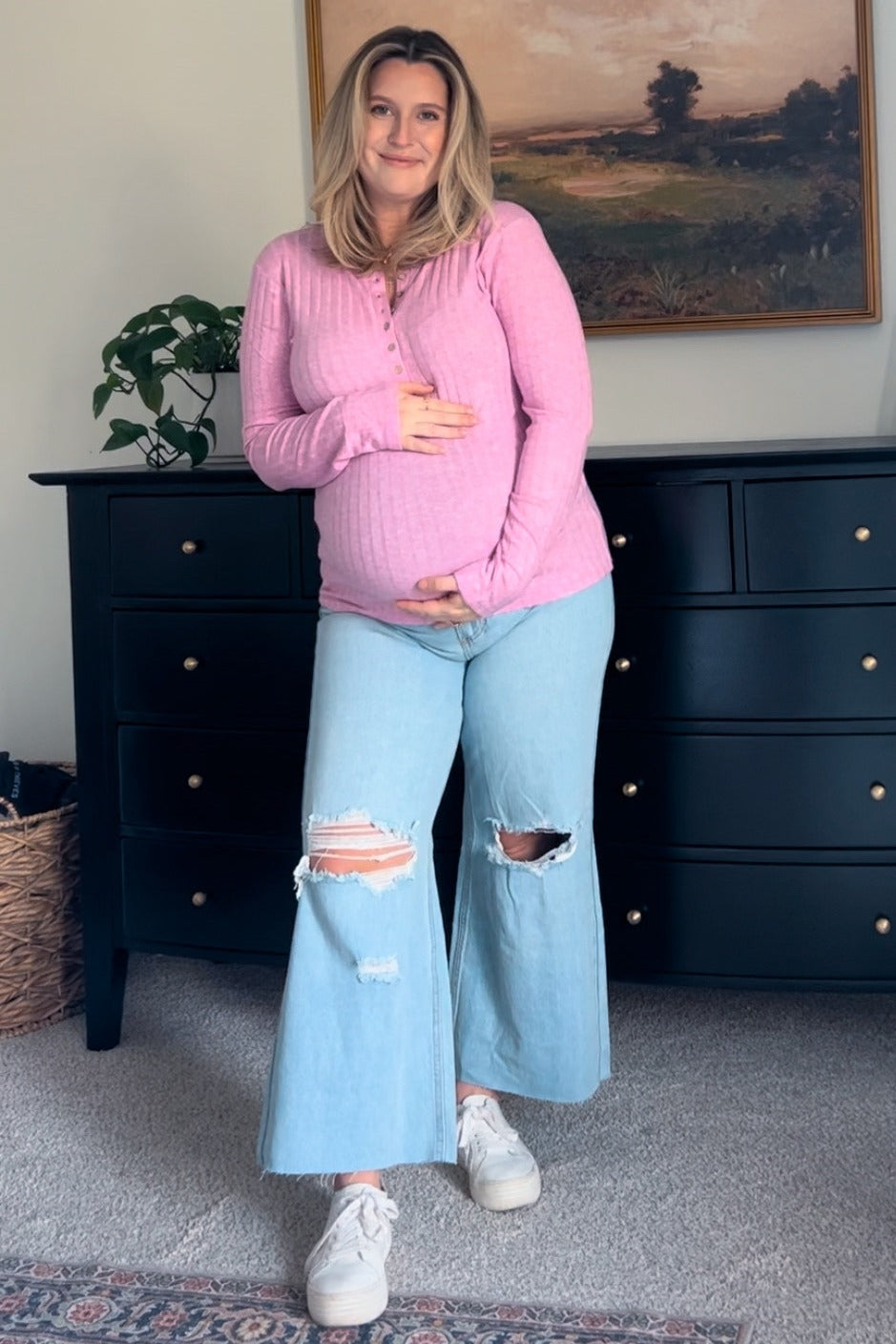 woman wearing a light pink long sleeve maternity top and distressed wide-leg maternity jeans, holding her baby bump