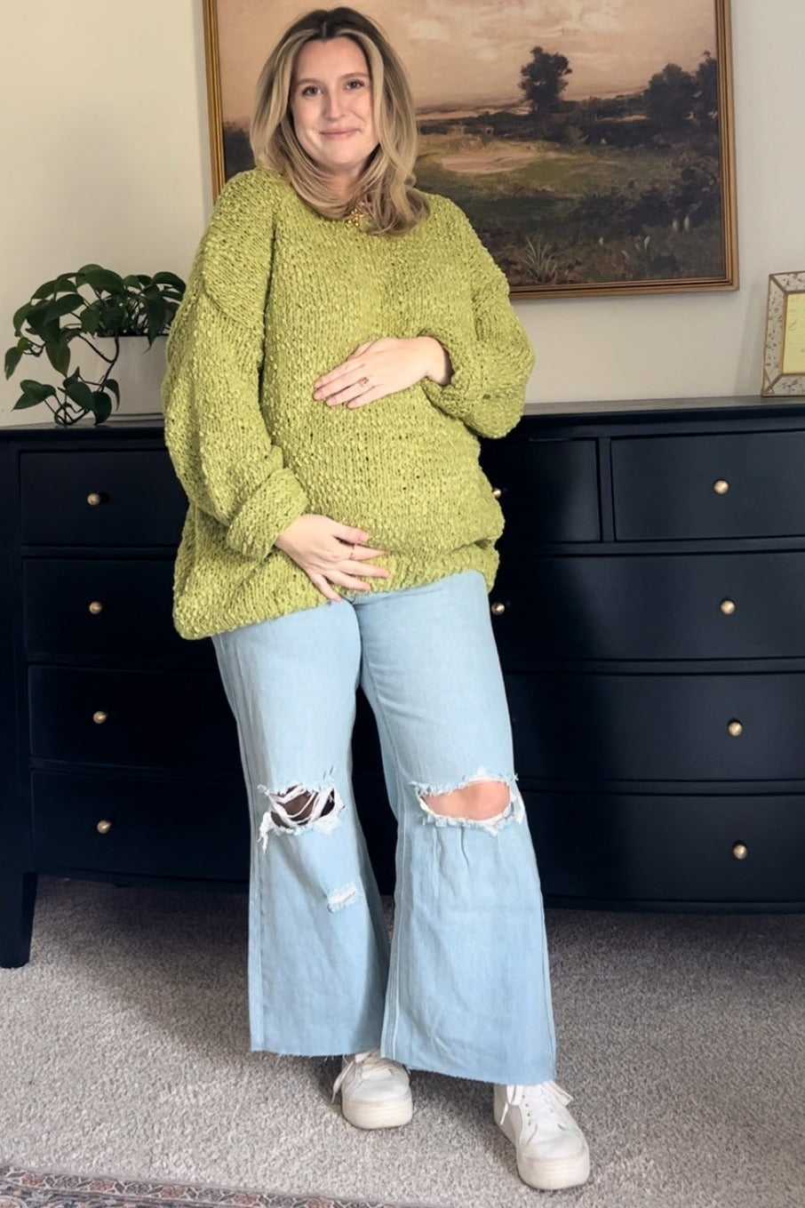 woman wearing a chunky green maternity sweater and distressed wide-leg jeans, cradling her baby bump