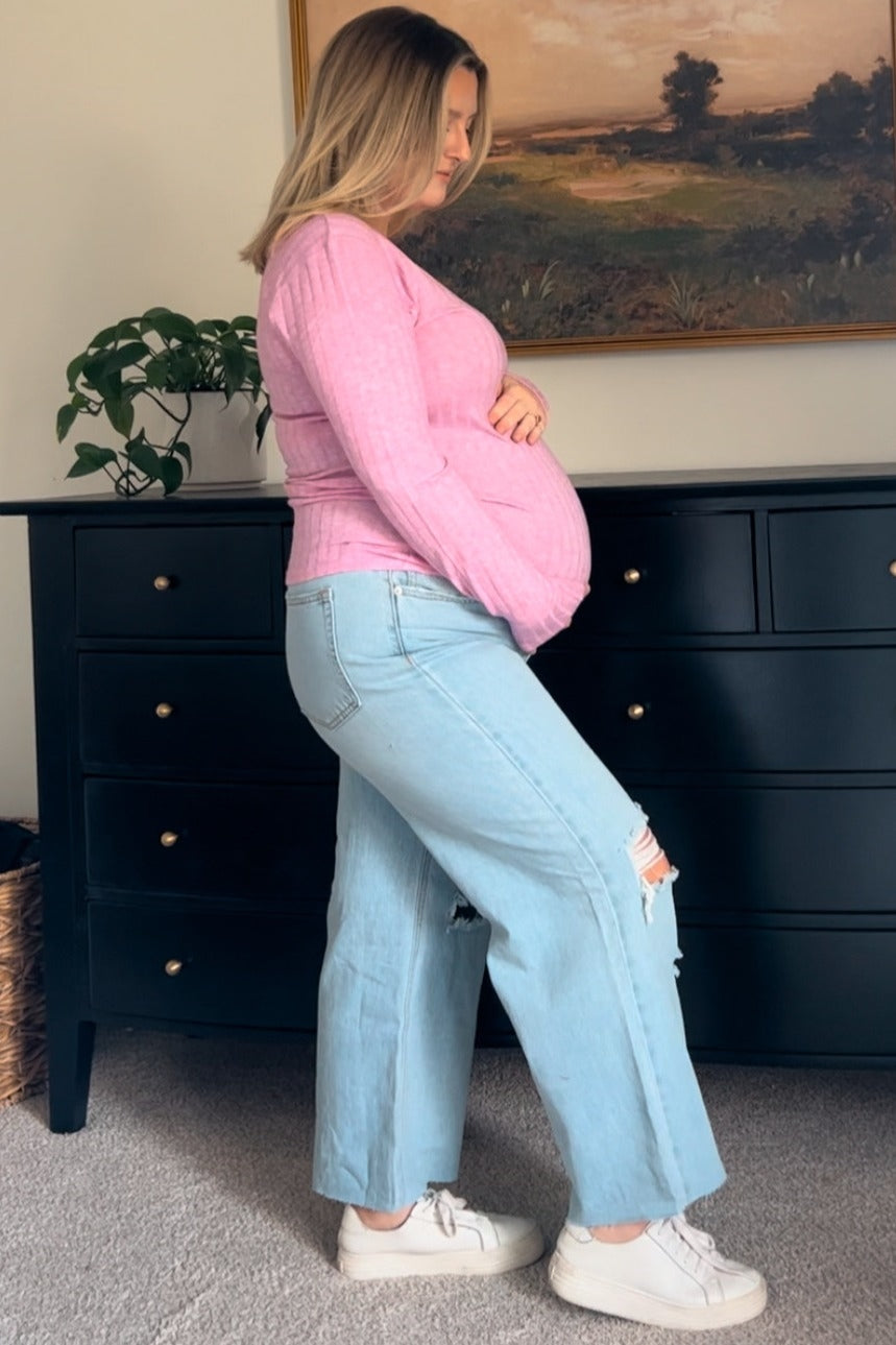 woman wearing a pink long sleeve maternity top and light blue distressed maternity jeans, showcasing her baby bump