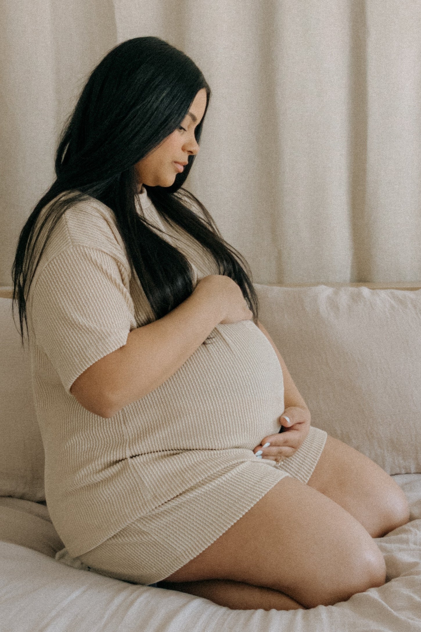 woman wearing a beige ribbed short sleeve maternity dress and holding her baby bump while sitting on a couch