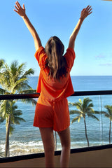 woman wearing an orange two-piece lounge set, standing on a balcony with arms raised, facing ocean and palm trees