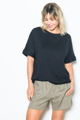 woman wearing a black oversized t-shirt and light beige shorts, smiling while posing against a white background