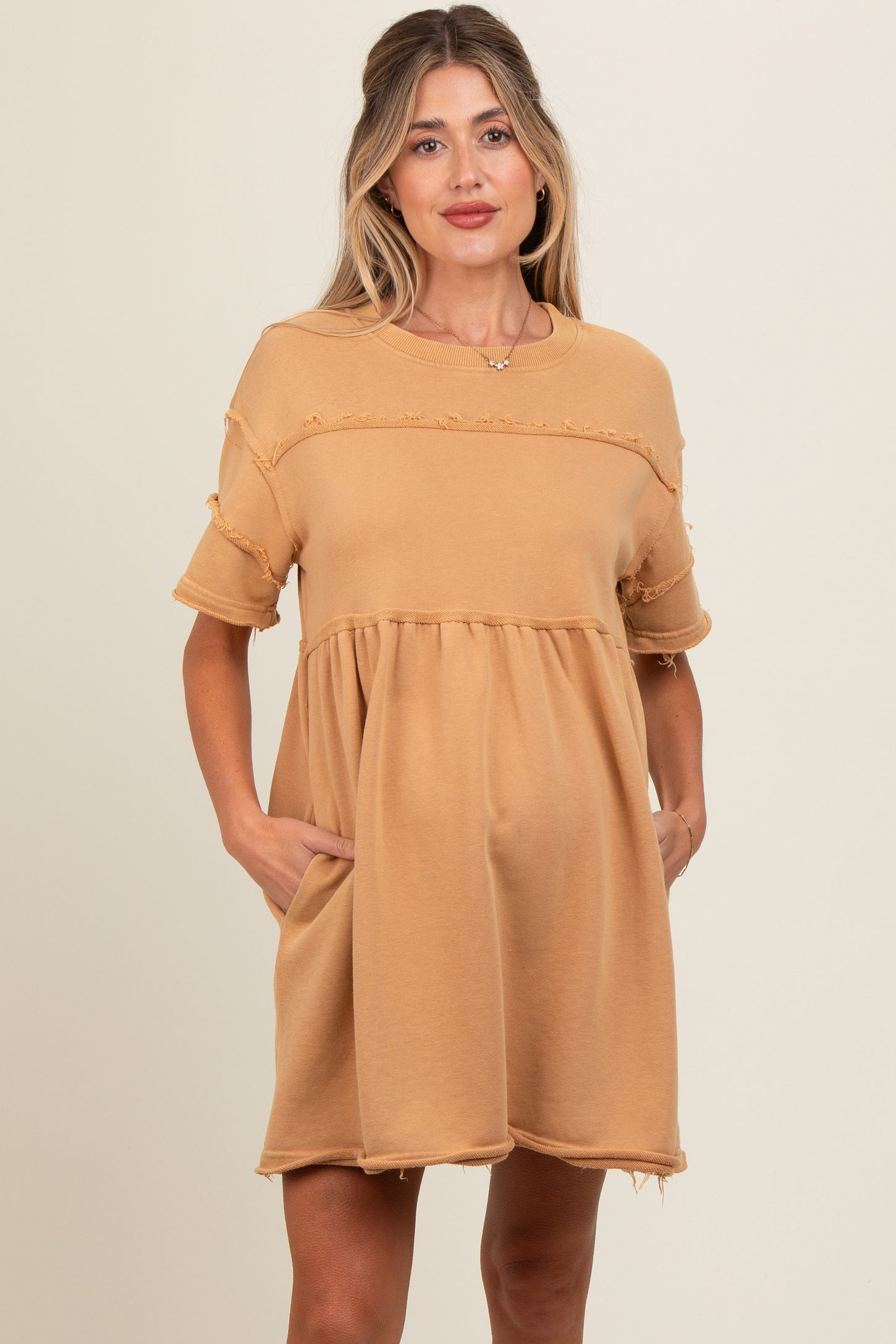 woman wearing a tan maternity dress with a tiered design and pockets, featuring distressed details and short sleeves