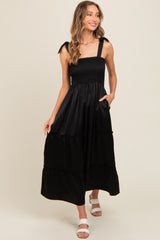 woman wearing a black smocked maxi dress with ruffled tiers and shoulder ties, standing and smiling