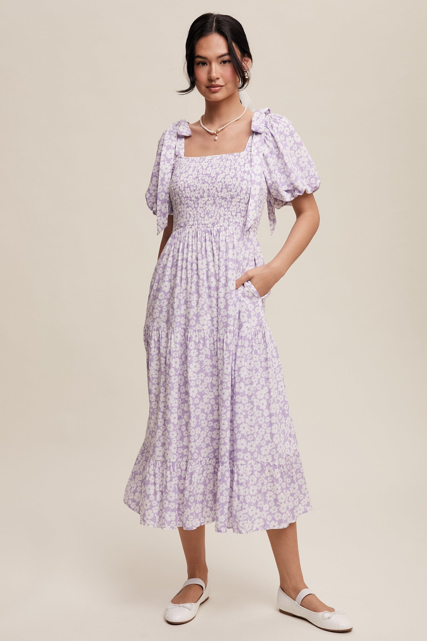 woman wearing a lavender floral print midi dress with puff sleeves and a smocked bodice, standing with one hand in a pocket