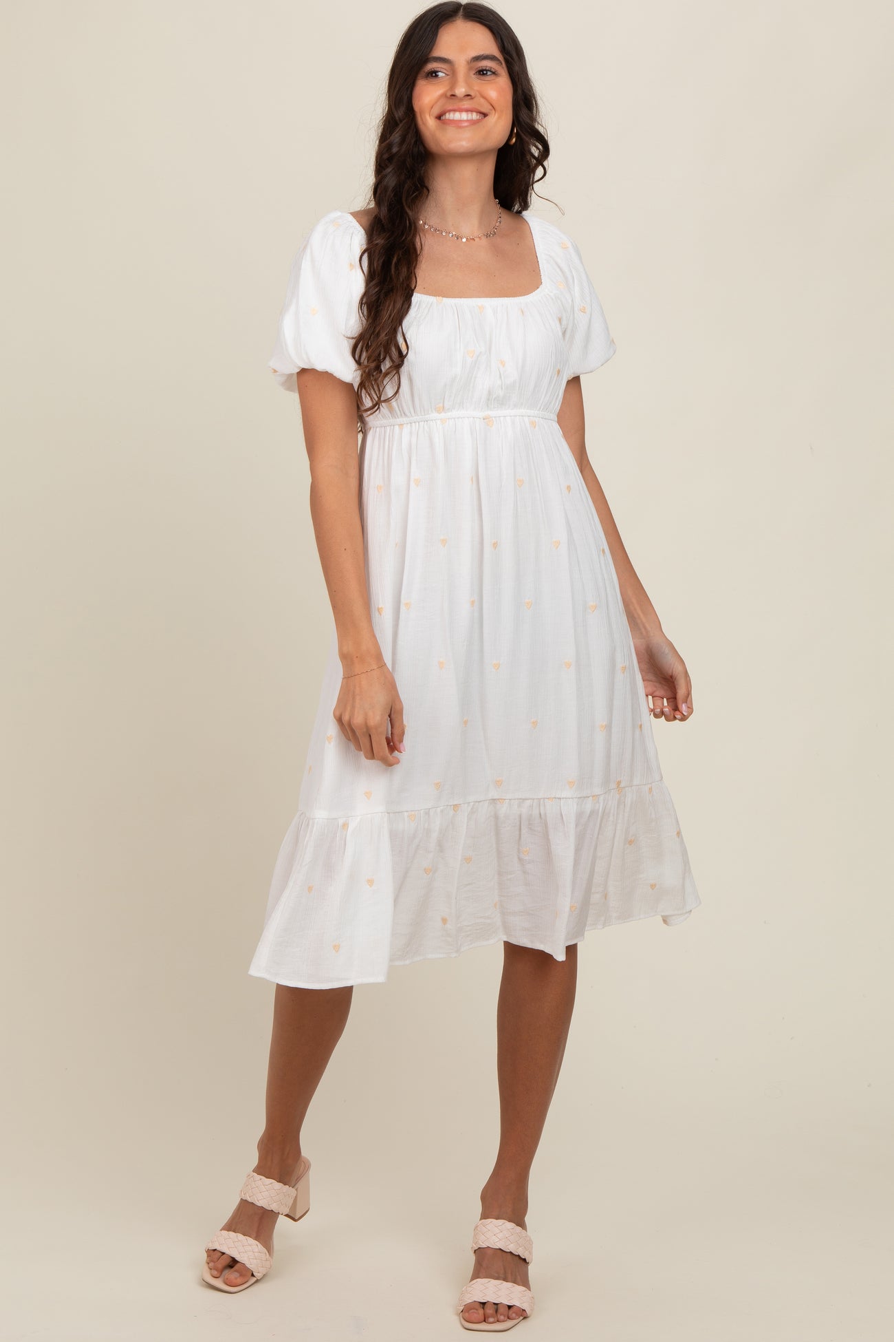 woman wearing a white puff sleeve midi dress with floral embroidery and a tiered hem, smiling and standing against a neutral background