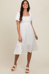 woman wearing a white puff sleeve midi dress with floral embroidery and a tiered hem, smiling and standing against a neutral background