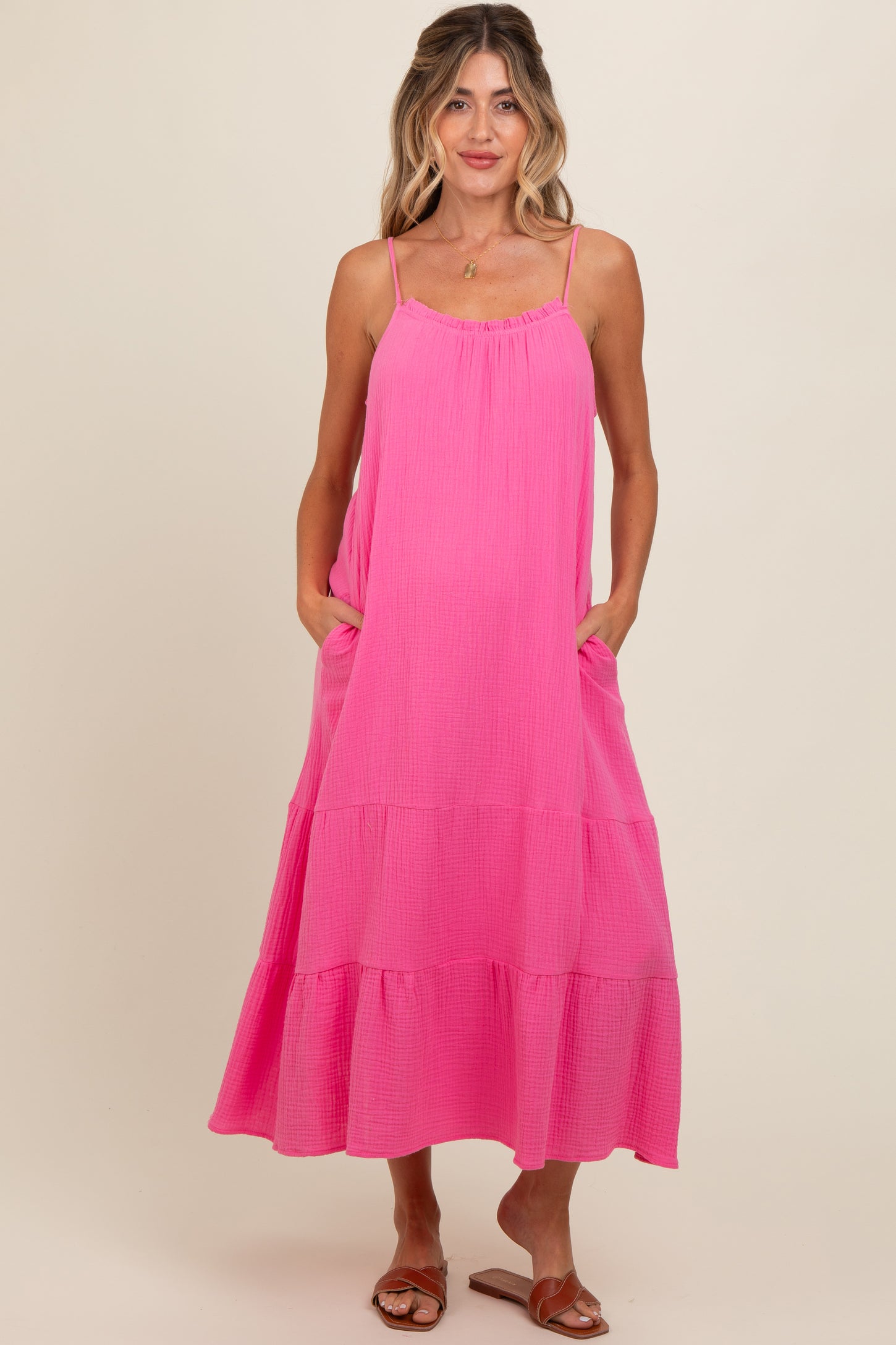 woman wearing a pink maternity dress with adjustable straps and tiered design, standing with hands in pockets