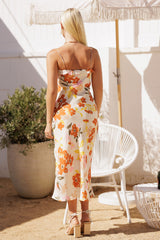 woman wearing a white floral midi dress with orange and pink flowers, seen from the back, standing outdoors by a plant