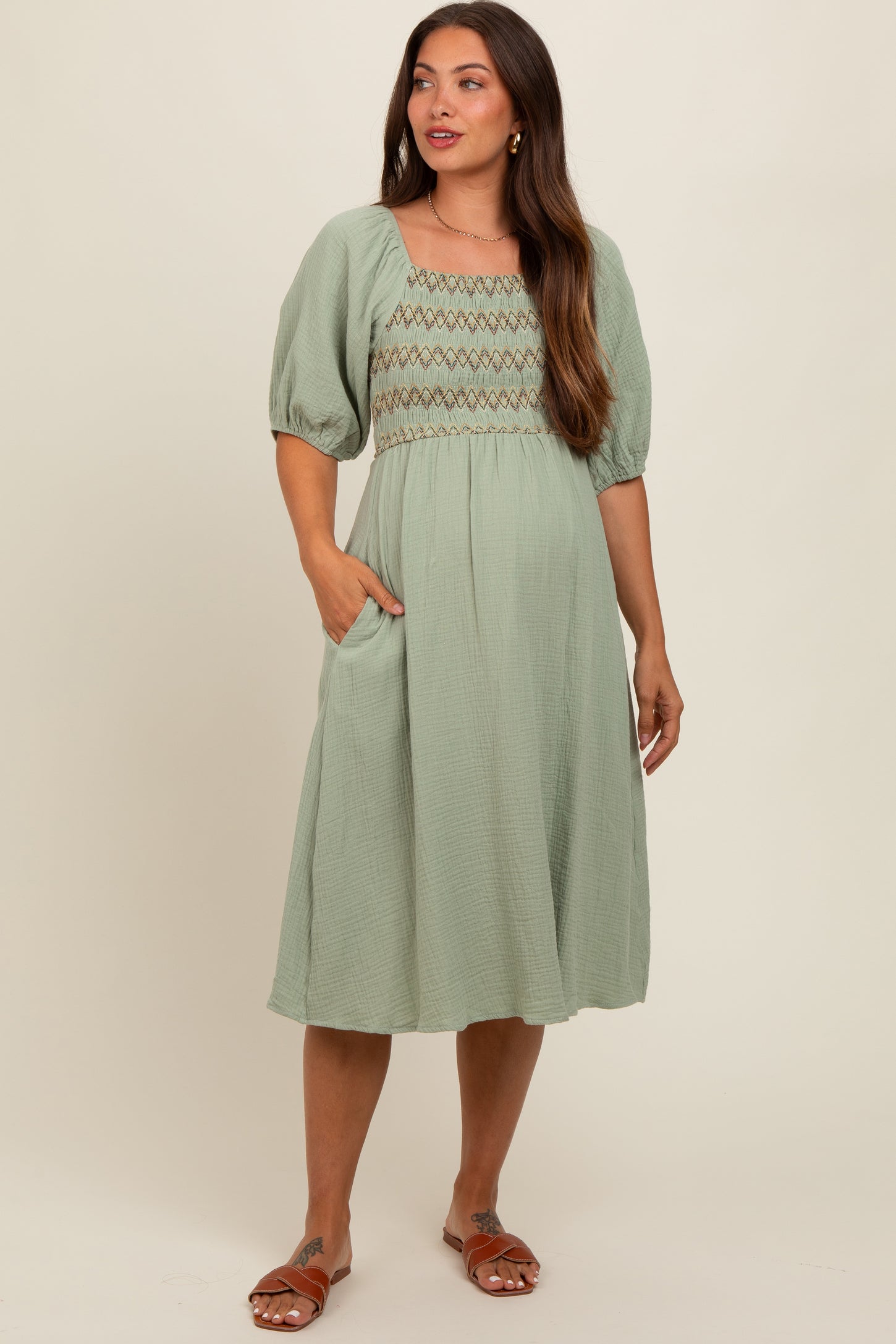 woman wearing a sage green maternity dress with puff sleeves and smocked bodice, posing with one hand in pocket