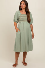 woman wearing a sage green maternity dress with puff sleeves and smocked bodice, posing with one hand in pocket