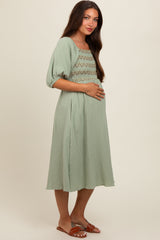 woman wearing a sage green short sleeve maternity dress with smocking detail and holding her baby bump