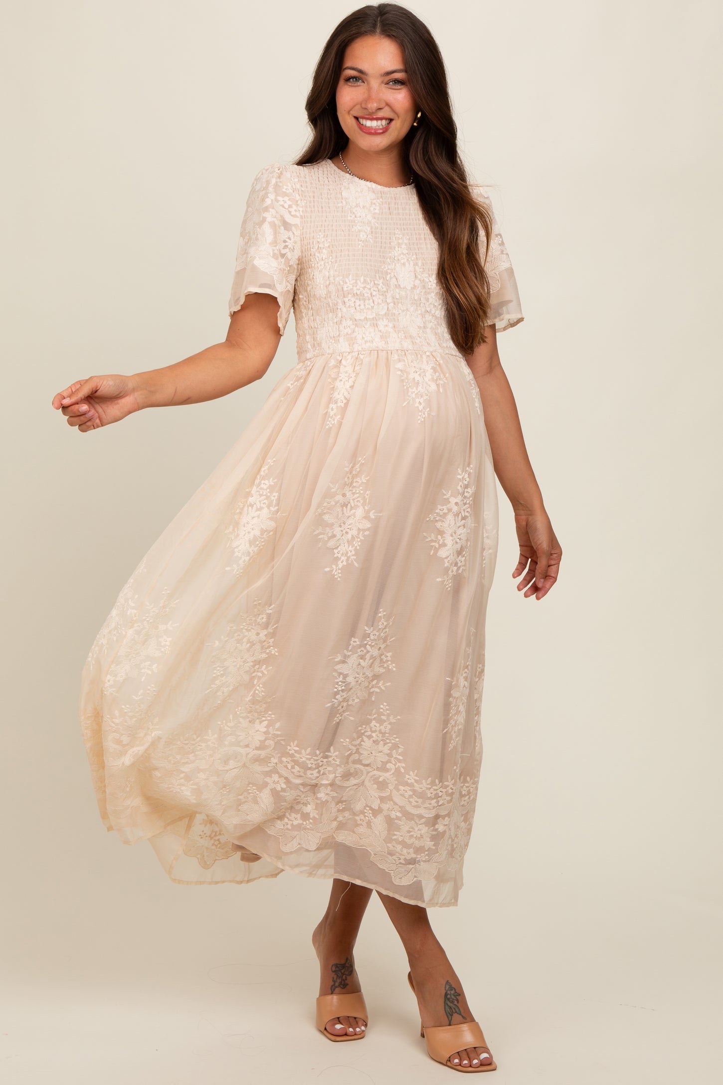 woman wearing a beige lace maternity dress with short sleeves and a flowing skirt, showcasing her baby bump