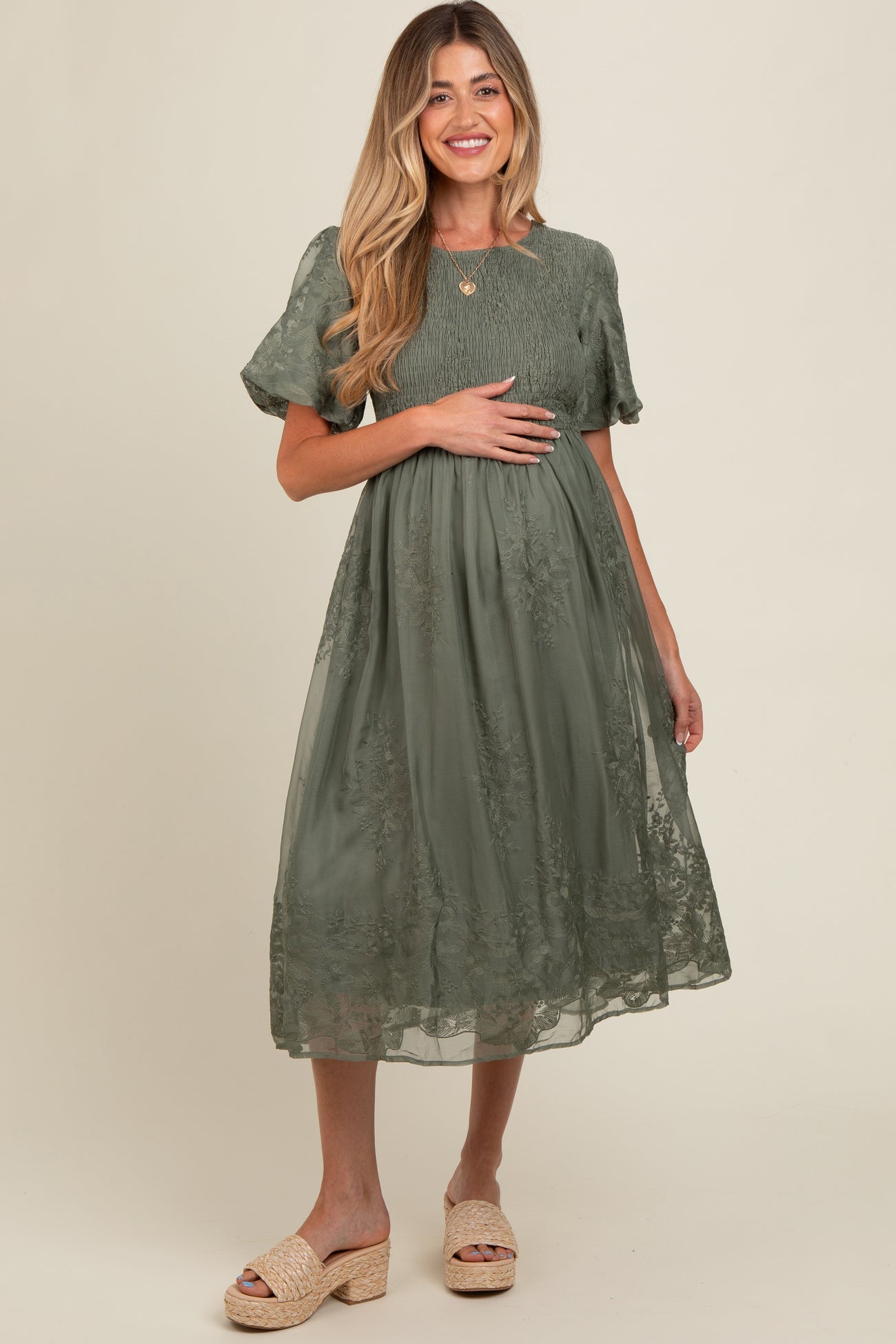 woman wearing a sage green maternity dress with embroidered details, holding her baby bump