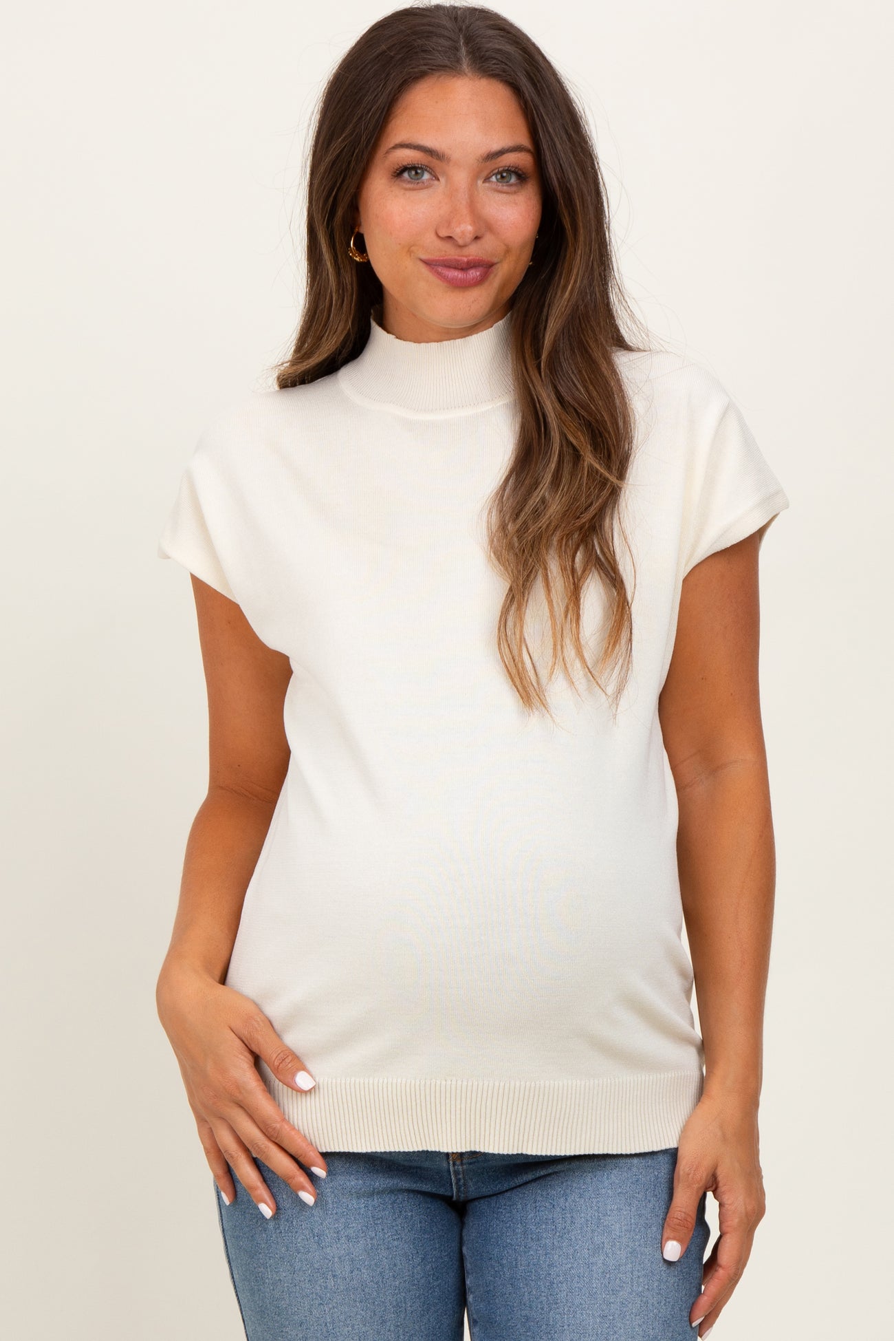 woman wearing a light beige maternity top with a mock neck and cap sleeves, showing her baby bump