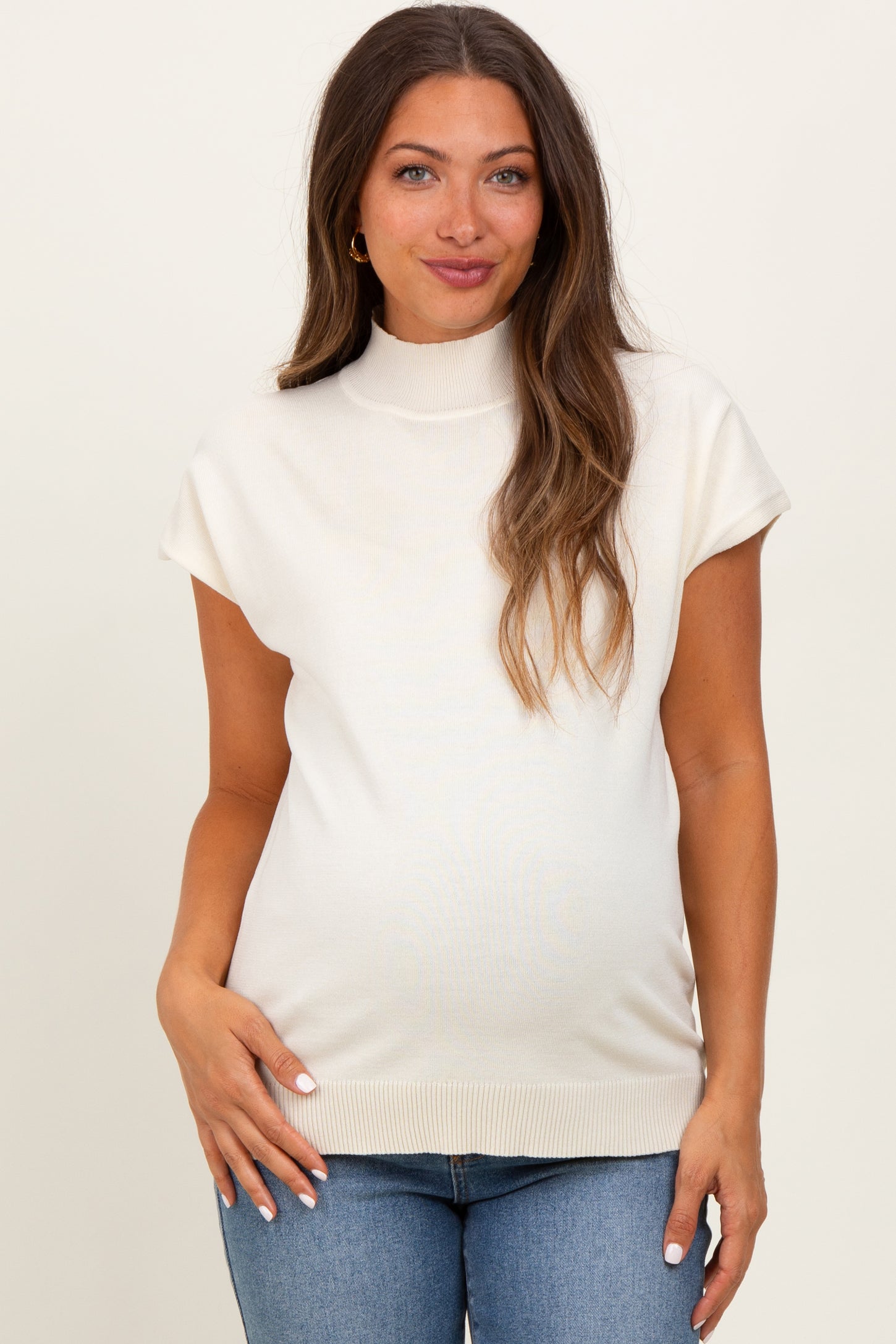 woman wearing a light beige maternity top with a mock neck and cap sleeves, showing her baby bump