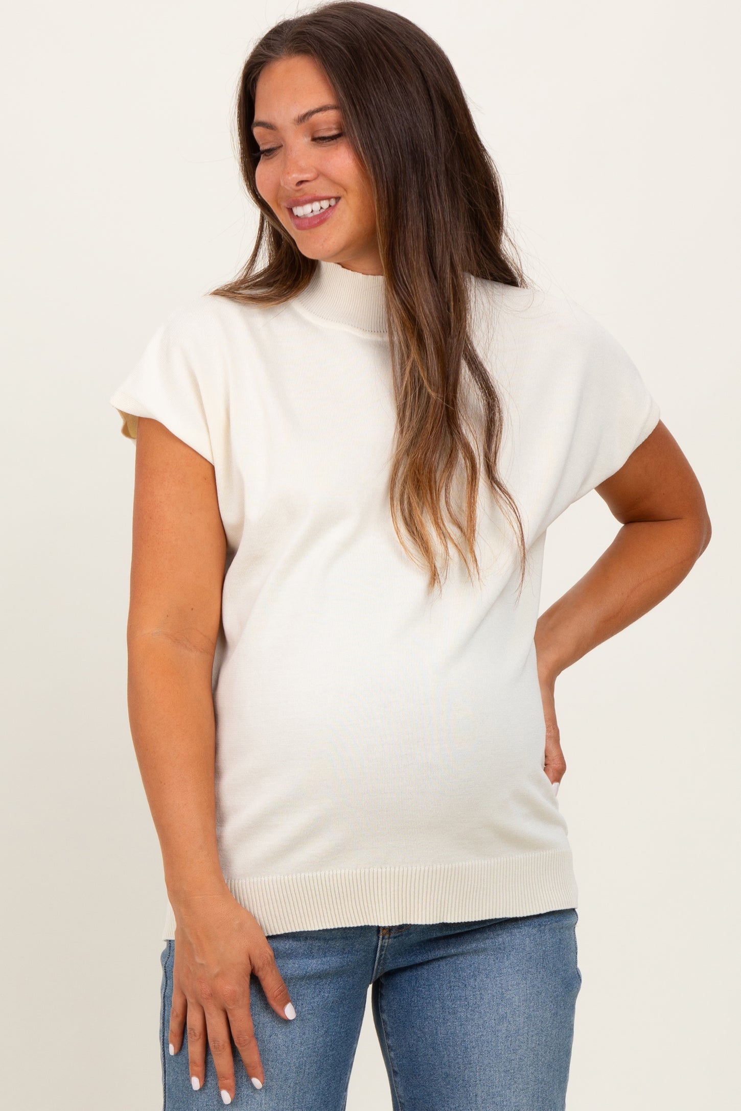 woman wearing a cream-colored short sleeve maternity top and blue jeans, showcasing her baby bump