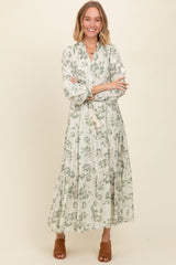 woman wearing a long sleeve white maxi dress with green floral print and a tiered skirt, standing with arms crossed