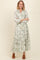 woman wearing a long sleeve white maxi dress with green floral print and a tiered skirt, standing with arms crossed