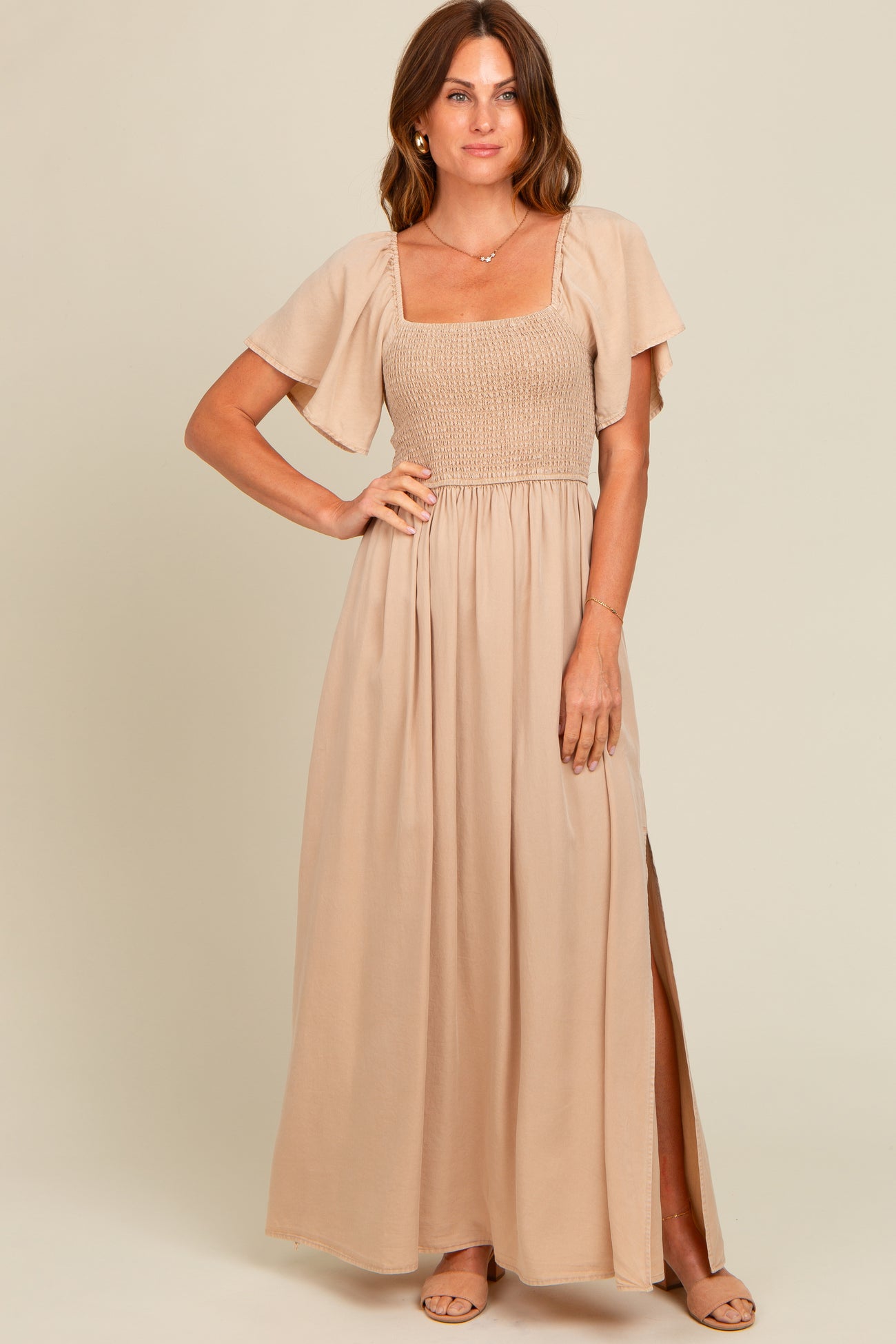 woman wearing a beige maxi dress with a smocked bodice and short flutter sleeves, showing a side slit