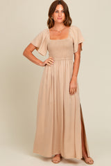 woman wearing a beige maxi dress with a smocked bodice and short flutter sleeves, showing a side slit