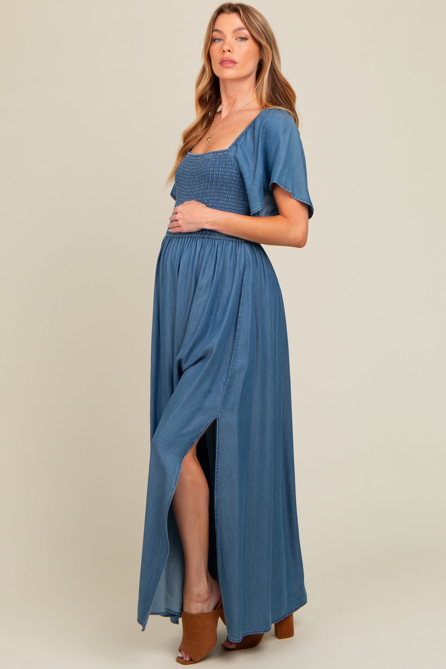 woman wearing a blue maternity maxi dress with a smocked bodice and side slit, holding her baby bump
