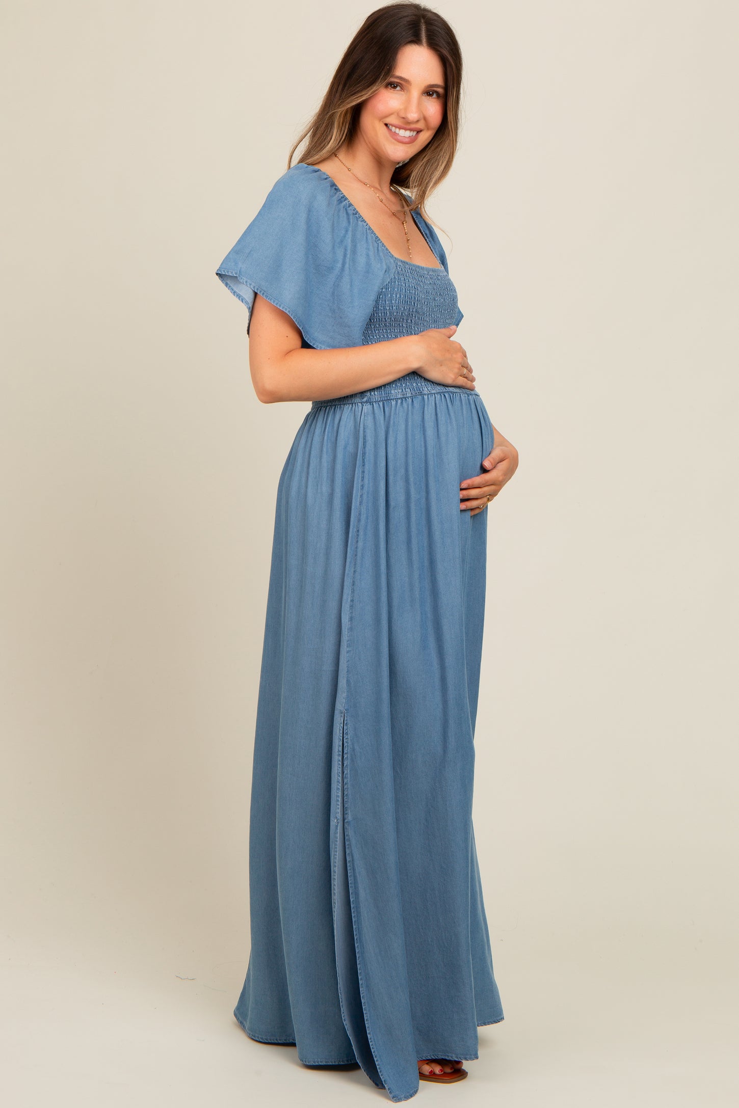 woman wearing a light blue maternity maxi dress with smocked bodice and flutter sleeves, gently cradling her baby bump