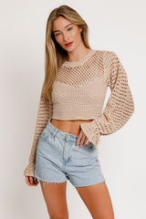 woman wearing a beige crocheted crop top with long bell sleeves and high-waisted denim shorts