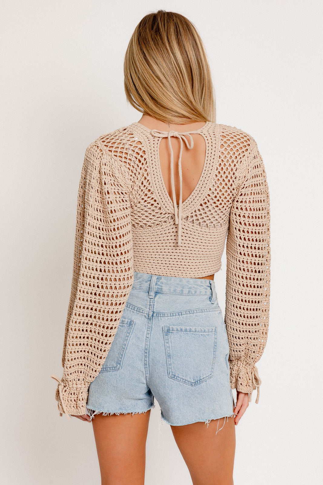 woman wearing a beige crochet top with long sleeves and back tie detail, paired with light blue denim shorts