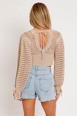 woman wearing a beige crochet top with long sleeves and back tie detail, paired with light blue denim shorts