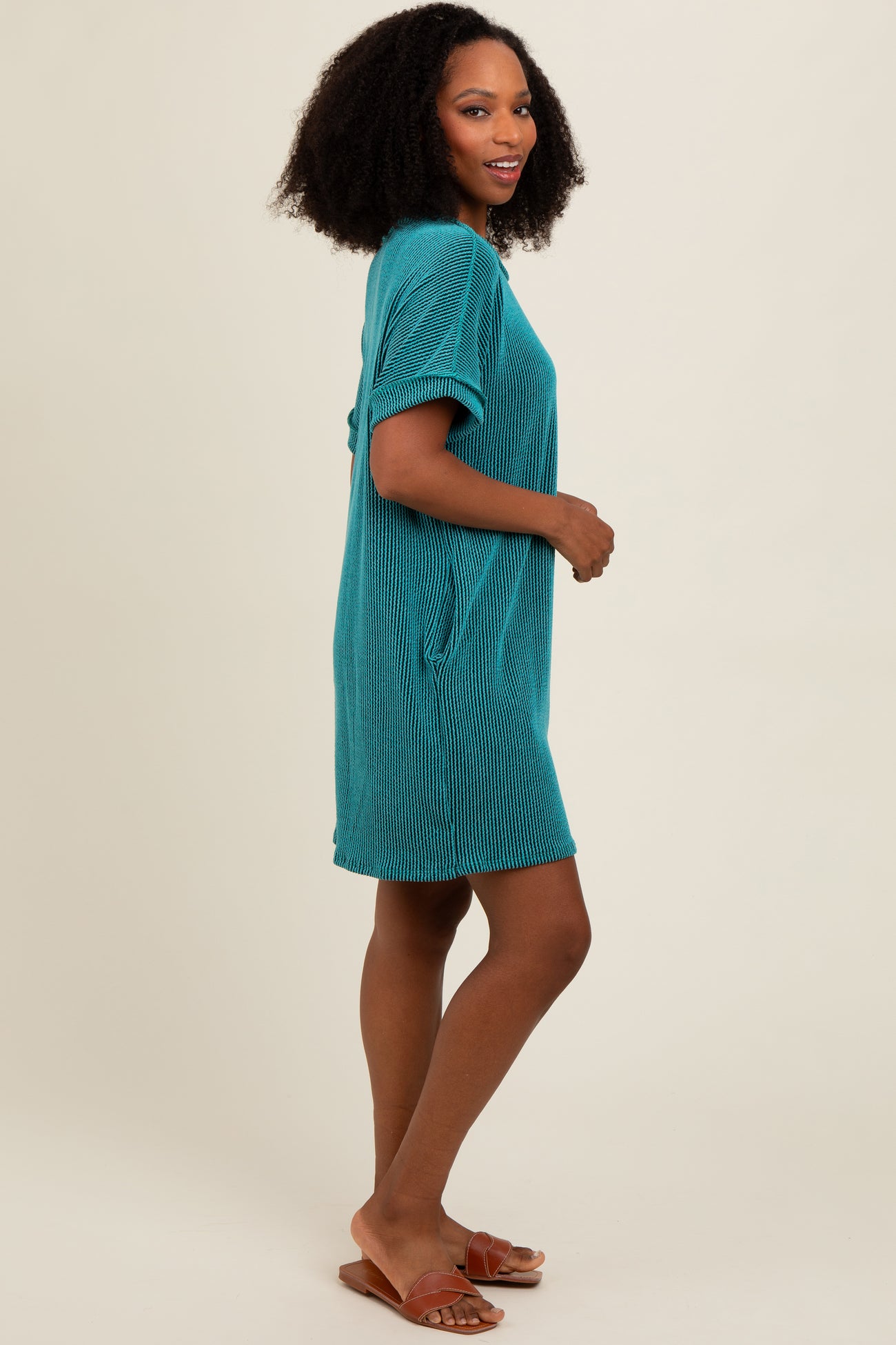 woman wearing a teal short sleeve striped dress with side pockets and sandals, standing sideways with a slight smile