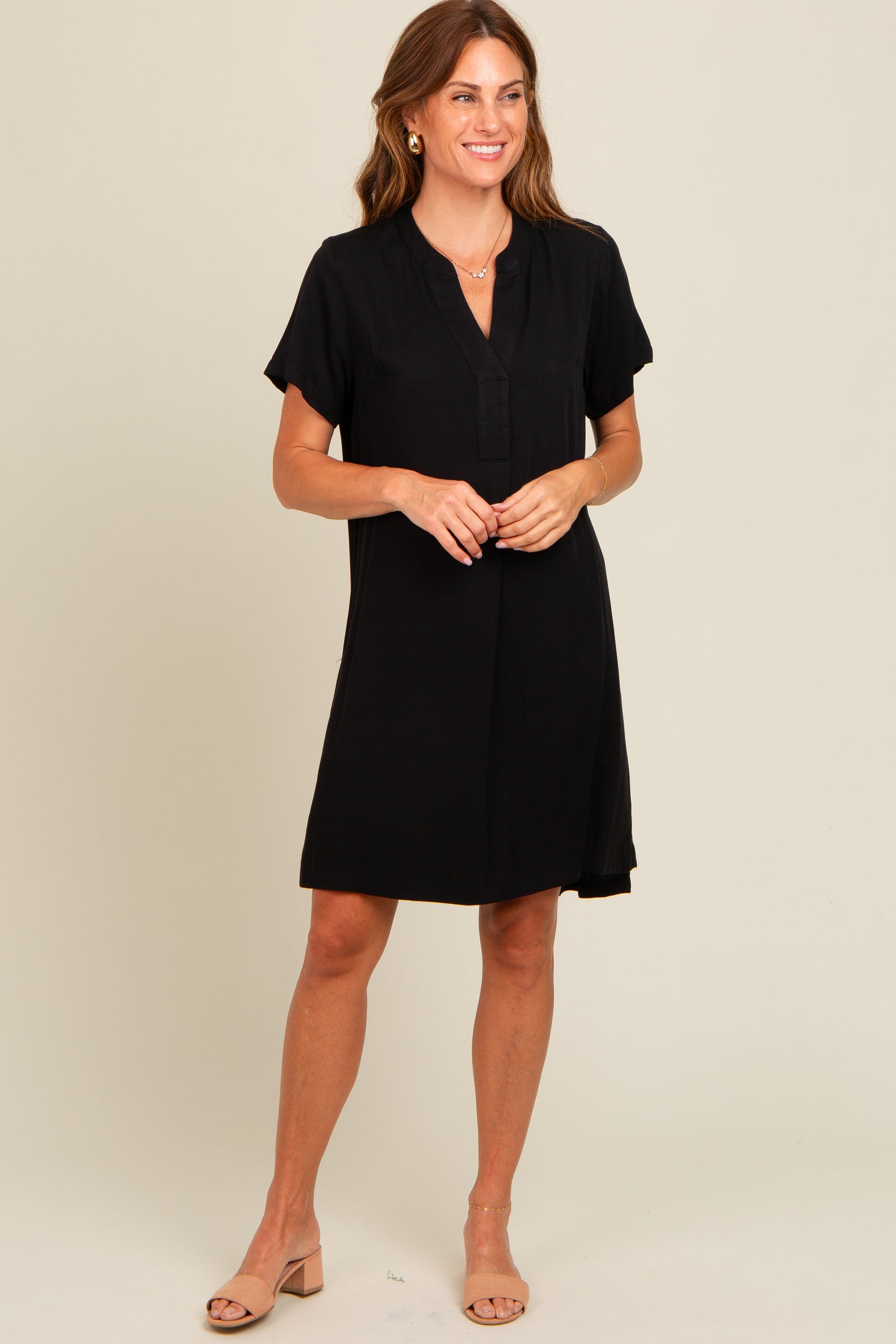 woman wearing a black short sleeve shift dress with a v-neck and button detail, styled with nude sandals