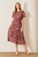 woman wearing a burgundy floral pleated midi dress with short sleeves and a tie waist, standing against a white wall