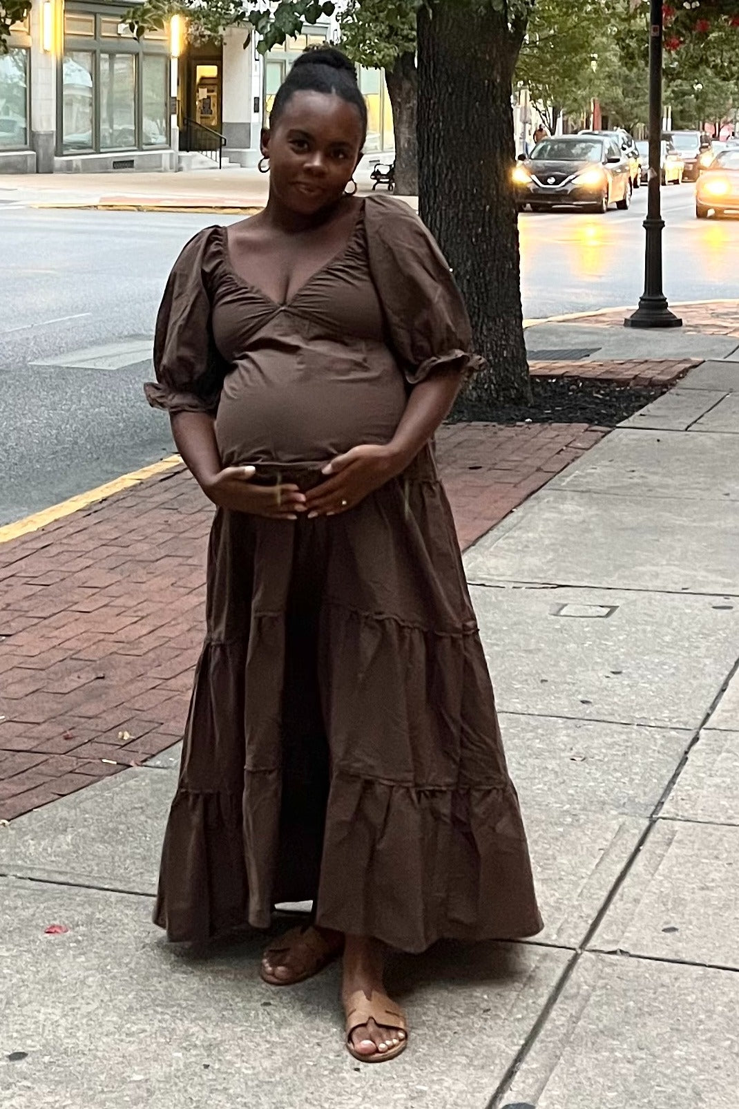 woman wearing a brown maternity maxi dress with puff sleeves and a tiered skirt, holding her baby bump outdoors
