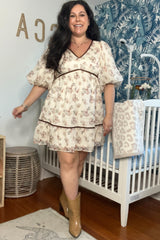 woman wearing a cream floral maternity dress with puff sleeves and a tiered design, standing beside a crib