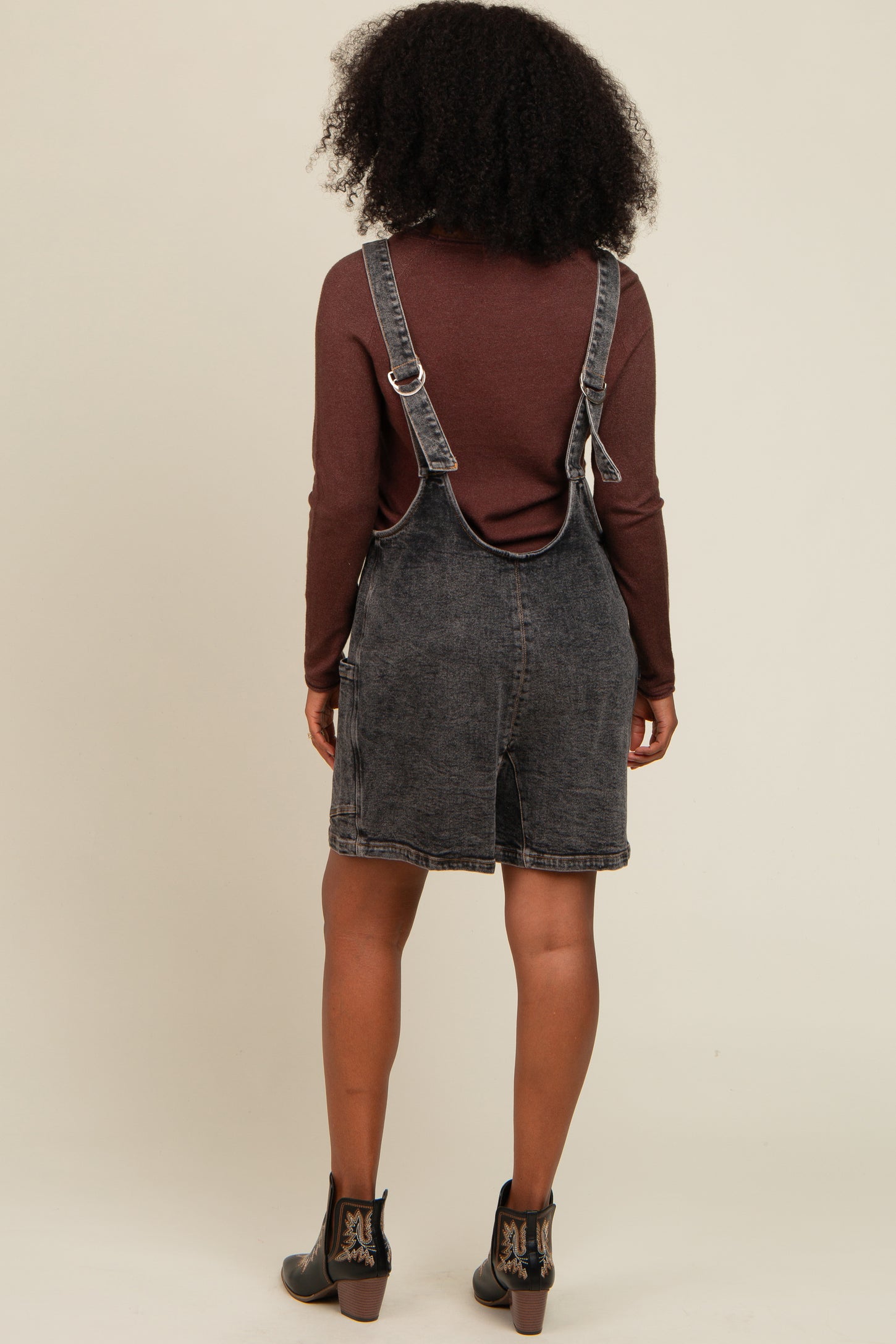 woman wearing a dark wash denim overall dress with a brown long sleeve top and black ankle boots from the back